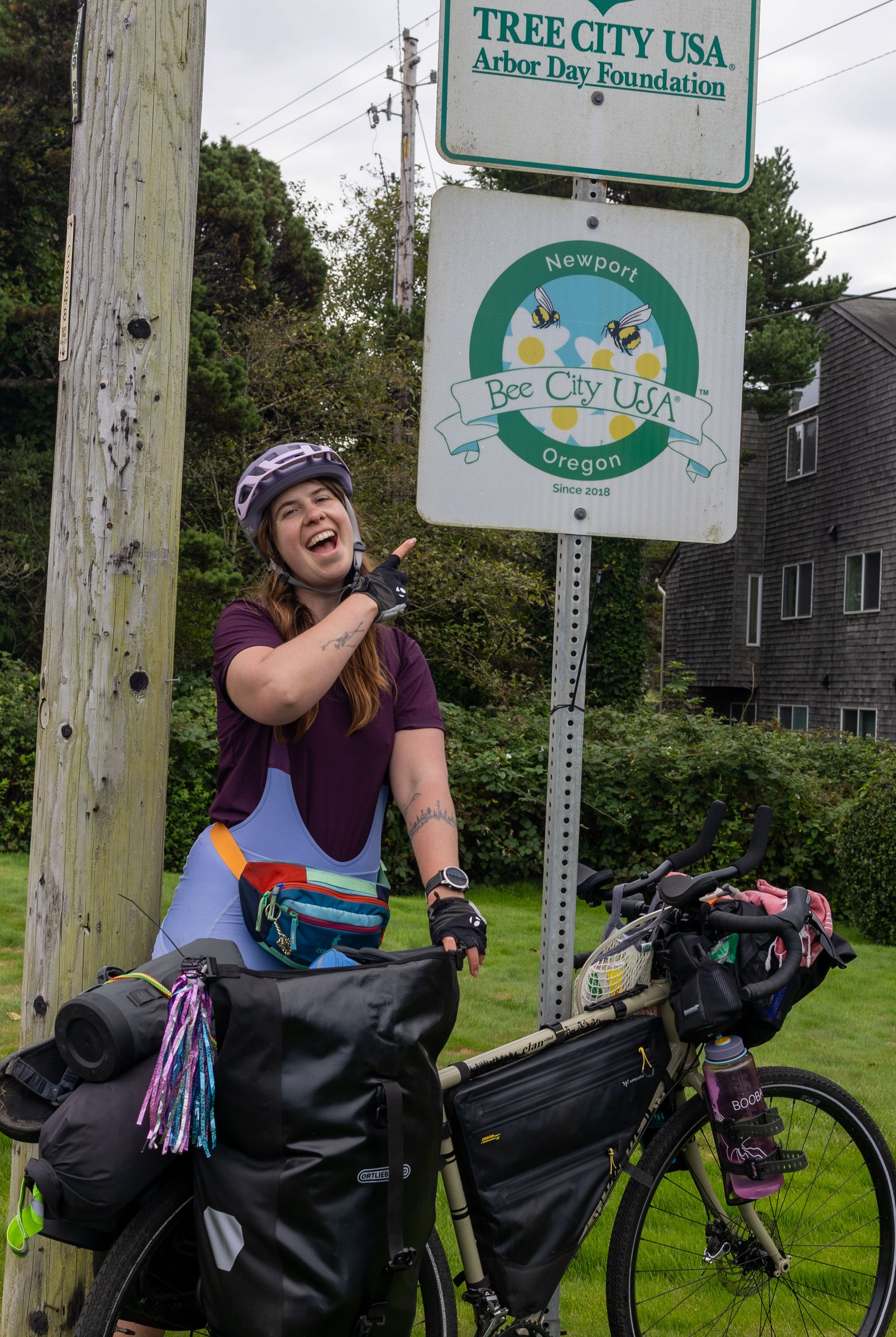 Bailey pointing to the Bee City USA sign in Newport, Oregon, beside her loaded touring bike