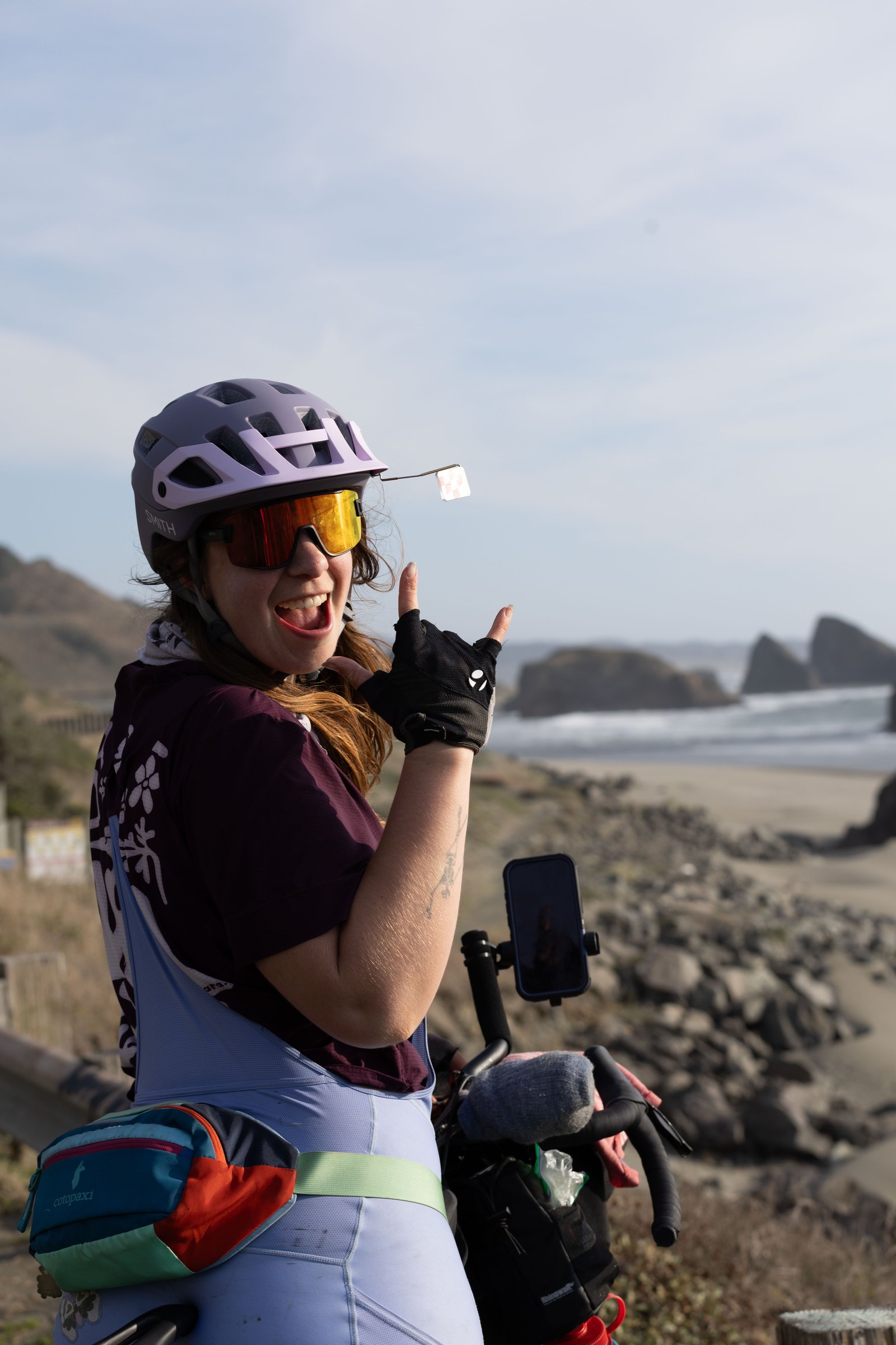 Bailey making a shaka sign beside her loaded bike on the coast